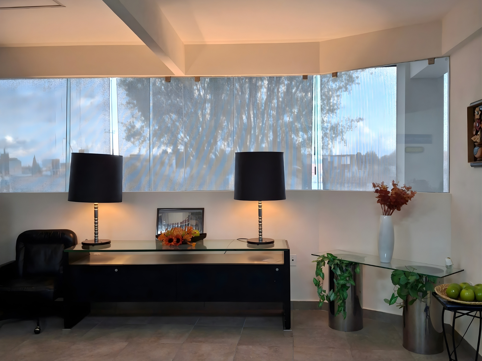 Sala de espera con consola oscura, dos lámparas de mesa con pantallas negras simétricas y ventanales grandes con vista al exterior difuminada.